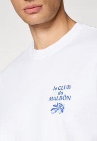 Hvid bomulds t-shirt med blå trykt tekst "le CLUB du MALBÔN" og et bladdesign, rund halsudskæring og korte ærmer.