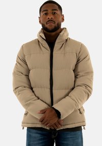 Beige puffjacka med ribbad textur, framdragkedja, hög krage och sidofickor. Bärs över en mörk topp, med en avslappnad passform.