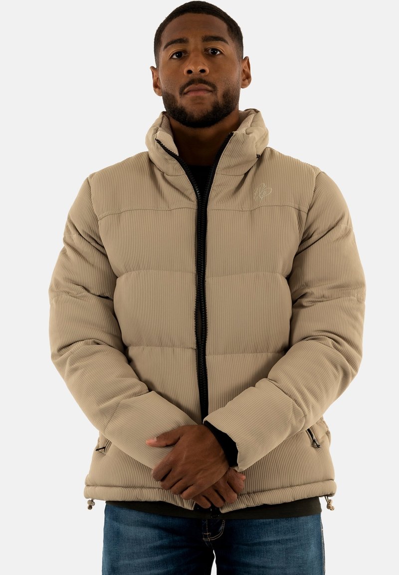 Beige puffjacka med ribbad textur, framdragkedja, hög krage och sidofickor. Bärs över en mörk topp, med en avslappnad passform.