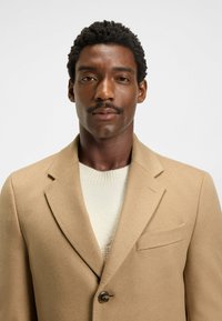 Cappotto di lana beige con colletto a rever, chiusura a un bottone e taschino sul petto, indossato sopra un maglione color crema. Tessuto con texture visibile.