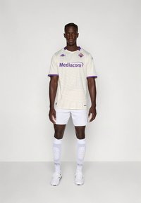 Baltas futbolo marškinėlis su purpuriniais akcentais, tekstūruotu raštu, su „Mediacom“ logotipu, derinamas su baltais šortais ir kelio aukščio kojinėmis.