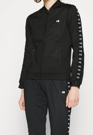 Chaqueta negra con cremallera y pantalones a juego con franjas repetidas de logotipos blancos a lo largo de las mangas y las piernas del pantalón en una persona de pie.