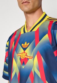 Adidas dres s krátkým rukávem má vícebarevný geometrický design v červené, žluté a modré barvě. Obsahuje vyšitá loga na hrudi.