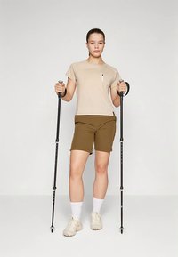 T-shirt beige avec une poche, short marron, chaussettes blanches et baskets claires ; tenant des bâtons de trekking noirs, debout devant un fond neutre.