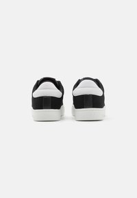 Fekete vászon edzőcipők fehér bőr kiegészítőkkel, texturált fekete sarkú panellel és gumitalppal. A hátsó nézet minimalista kialakítást mutat.
