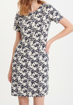 Frau trägt ein kurzärmeliges, knielanges beiges Kleid mit einem marineblauen Blumenmuster, steht mit einer Hand in der Hüfte.