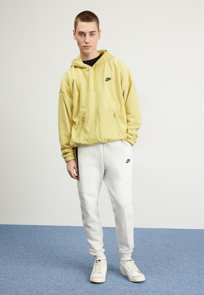 Sudadera de felpa amarilla con un bolsillo frontal y un logo negro, combinada con pantalones de chándal gris claro que tienen un logo negro. Zapatillas blancas completan el look.