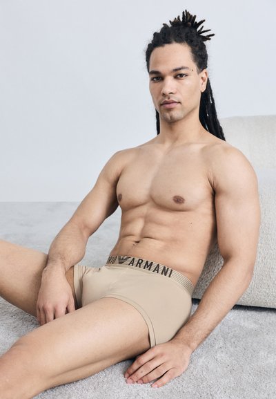 Άνδρας με μακριά dreadlocks ξαπλωμένος σε χαλί, φορώντας μπεζ εσώρουχα Armani, εμφανίζοντας μυώδη κορμό και χαλαρή στάση.