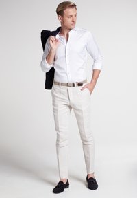 Camicetta bianca con bottoni, pantaloni slim-fit beige chiaro, cintura marrone e mocassini neri. L'uomo tiene un blazer nero sopra la spalla.