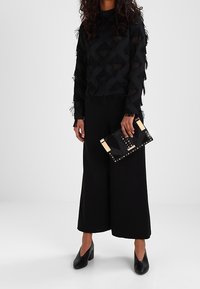 Haut noir à manches longues texturé avec des détails en frange, associé à un pantalon noir à jambe large, tenant une pochette noire avec des accents dorés et des clous.