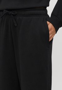 Mano nella tasca dei pantaloni sportivi neri con vita regolabile, indossati con una maglia nera a maniche lunghe.