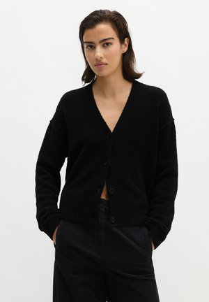 Junge Frau mit dunklen Haaren trägt eine schwarze, geknöpfte Strickjacke und schwarze Hose, steht mit Händen in den Taschen vor einem schlichten Hintergrund.