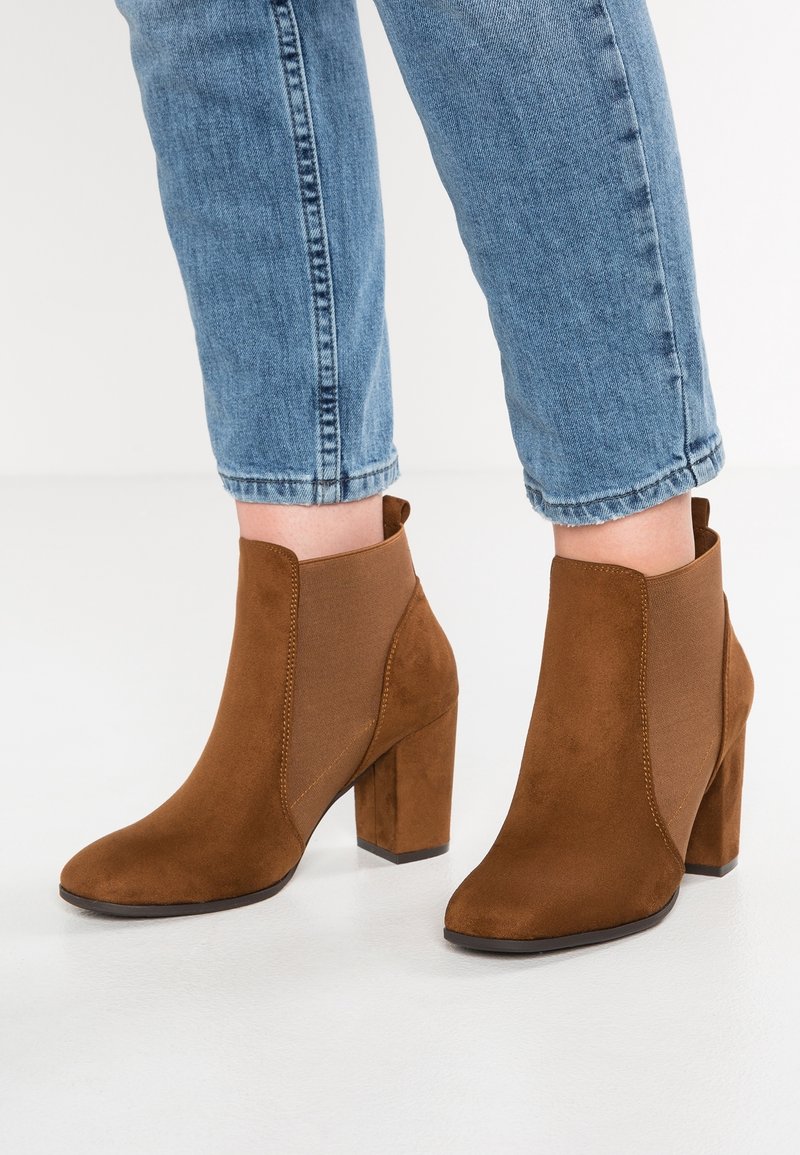 Bottes chevilles en daim marron avec talons carrés portées avec un jean denim bleu retroussé sur fond blanc.