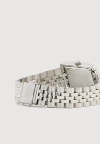 Sidabrinis metalinis pirštinis laikrodis su stačiakampiu korpusu, reguliuojamu grandinėlės apyrankiu ir tekstūruotu paviršiumi. Sagties užsegimas turi išgraviruotą logotipo detalę.