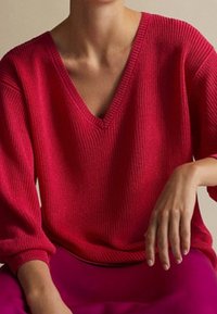 Personne portant un pull côtelé rouge vif à col en V avec des manches larges, reposant un bras sur son genou devant un fond neutre.