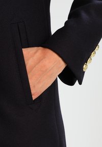 Marinblå kapp med ficka på ärmen, i strukturerad tyg med guldfärgad knappdetalj på mudden. Närbild av hand i fickan.