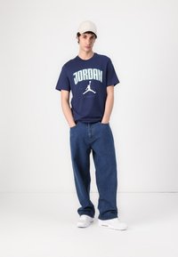 Mørkeblå bomulds t-shirt med "JORDAN" grafik og logo. Kombination med løstsiddende blå denim jeans og hvide sneakers. Hvid kasket.