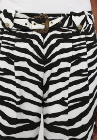 Melnā un baltā zebras svītrojuma bikses ar krokām, ar dekoratīvu zelta sprādzi un atbilstošām pogām jostasvietā.