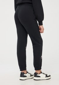 Pantaloni della tuta neri con polsini elasticizzati, dotati di accenti grafici a contrasto sui lati. Abbinati a sneakers bianche e nere.