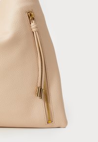Beige Lederhandtasche mit strukturierten Oberfläche, schräger Form und einem seitlichen Reißverschluss mit goldenen Beschlägen zum Verschließen.