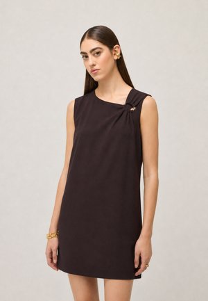 Donna con lunghi capelli scuri che indossa un vestito scuro senza maniche con un dettaglio ad anello sulla spalla e gioielli dorati a orecchio e polso.