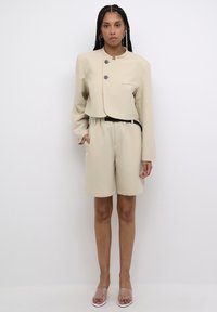 Beige cropped Jacke mit zwei schwarzen Knöpfen und Taschendetails; kombiniert mit knielangen Shorts, beide aus glattem Stoff. Das Modell trägt transparente Sandalen.