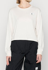 Long-sleeve, light beige t-shirt in soft fabric with a cropped design, featuring a black logo patch on the left chest.