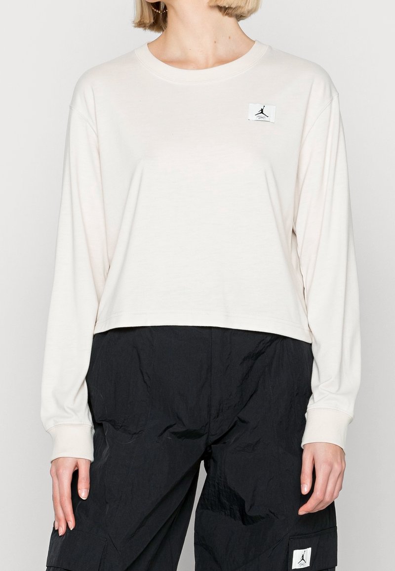 Long-sleeve, light beige t-shirt in soft fabric with a cropped design, featuring a black logo patch on the left chest.
