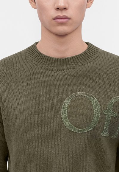 Homme portant un pull en maille vert olive avec un col côtelé haut et des lettres partiellement brodées "Of" sur la poitrine.