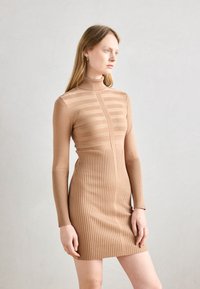 Robe en tricot beige avec un col montant, présentant des rayures horizontales en haut et des côtes verticales sur la jupe. Sa coupe ajustée met en valeur la forme.