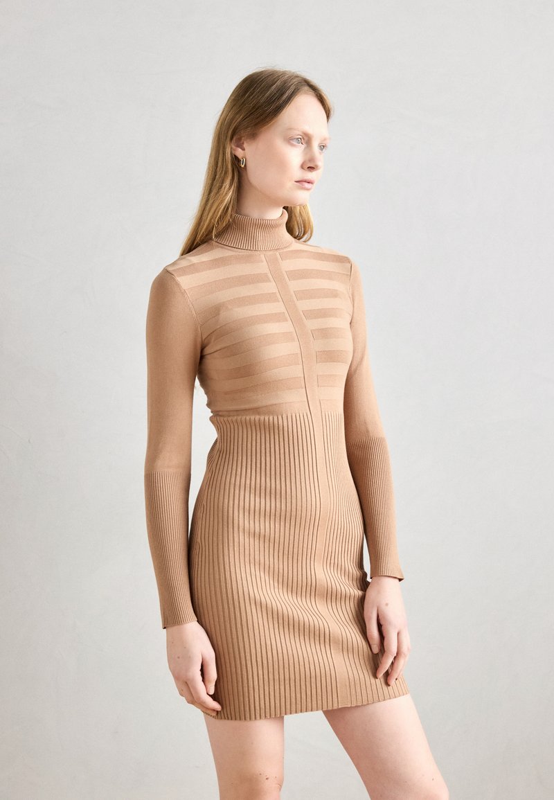 Robe en tricot beige avec un col montant, présentant des rayures horizontales en haut et des côtes verticales sur la jupe. Sa coupe ajustée met en valeur la forme.