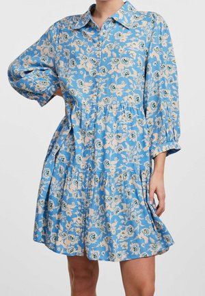 Femme portant une robe bleue à fleurs avec de longues manches bouffantes, un col et une jupe à étages, debout avec une main sur la hanche.