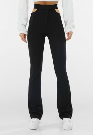 Vrouw draagt een zwarte broek met hoge taille en zijuitsnijdingen, gecombineerd met een wit cropped top met lange mouwen en witte sneakers.