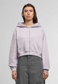 Lyselilla cropped hoodie med lynlås, store frontlommer og elastiske manchetter. Har et lille broderet logo på venstre brystområde.
