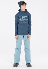 Felpa con cappuccio blu navy con tasca frontale, caratterizzata da una stampa grafica. Pantaloni da sci blu chiaro, stivali neri impermeabili. Sfondo bianco uniforme.