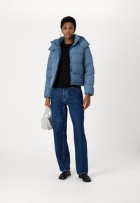 Giacca a vento azzurro chiaro con cappuccio, caratterizzata da una chiusura a zip e texture trapuntata. Indossata sopra una maglietta nera con jeans blu a vita alta.