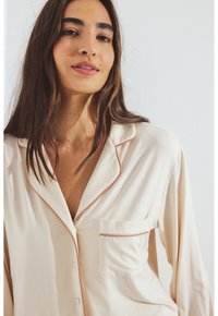 Beige satijnen pyjama top met een notch-kraag, contrasterende stiksels, borstzak en knoopsluiting. Zachte, gladde textuur.