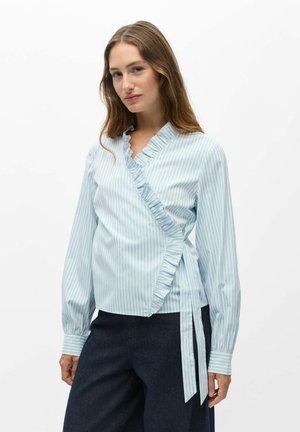 Femme portant un chemisier cache-cœur rayé bleu clair et blanc avec bordure à volants et pantalon foncé, posant devant un fond blanc uni.