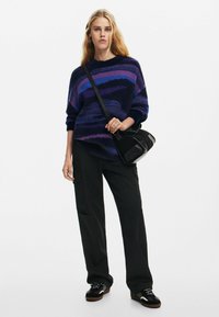 Oversized gebreide trui in marineblauw en paarse strepen, gecombineerd met zwarte cargo pants en een zwarte schoudertas, met een relaxte pasvorm en textuur.
