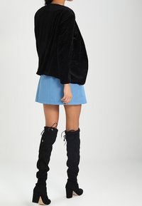 Haut oversize en velours noir à manches longues, associé à une mini-jupe bleue et des bottes cuissardes en suède noir à talon carré.