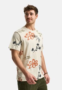 Camiseta de manga corta color beige claro con estampado floral en naranja, negro y verde. Presenta un cuello redondo y un corte holgado.