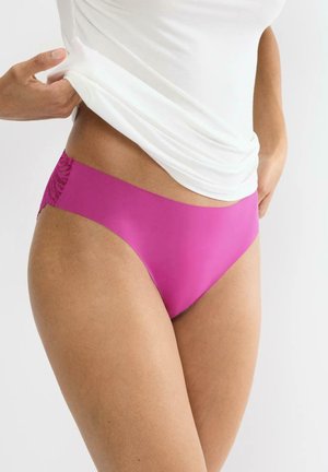 Kobieta ubrana w bezszwowy magentowy bikini i białą koszulę, podnosząca koszulę, aby pokazać gumkę w talii i koronkowe detale po bokach.