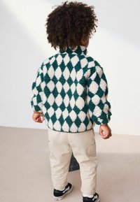 Veste en polaire à motif losange vert et blanc avec un col haut, associée à un pantalon cargo beige et des baskets noires.