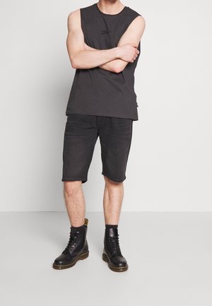 Hombre vestido con una camiseta de tirantes negra, pantalones cortos negros hasta la rodilla y botas negras con cordones, usando calcetas negras, de pie con los brazos cruzados.