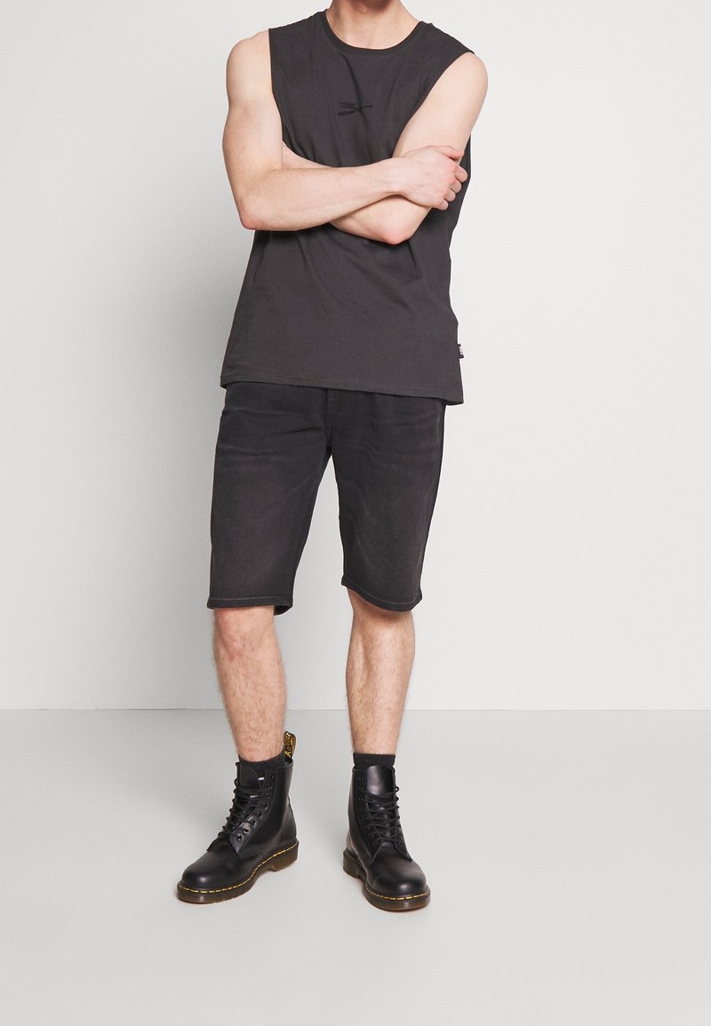 Man wearing black sleeveless shirt, black knee-length shorts, and black lace-up boots with black socks, standing with arms crossed.