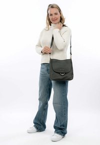Borsa in pelle grigia con pattina arrotondata, accento metallico circolare e tracolla regolabile. Una donna con un maglione crema e jeans blu tiene la borsa.
