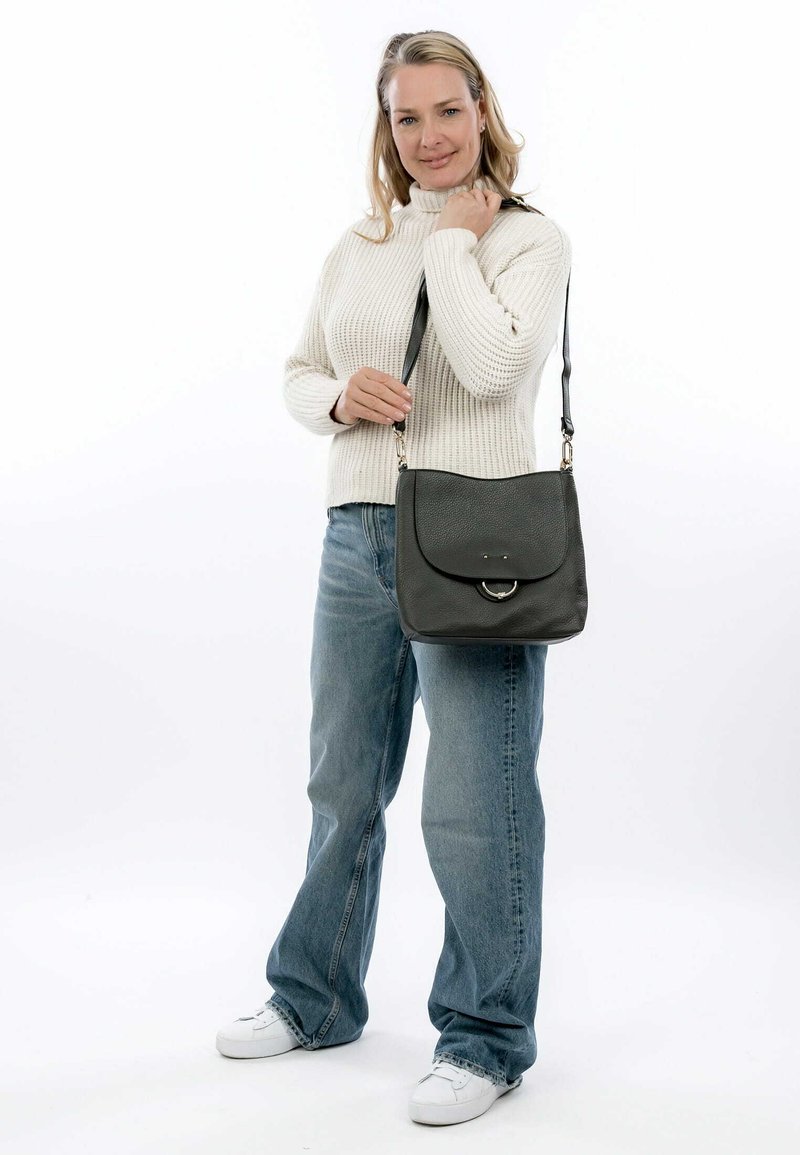 Borsa in pelle grigia con pattina arrotondata, accento metallico circolare e tracolla regolabile. Una donna con un maglione crema e jeans blu tiene la borsa.