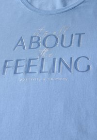 Camiseta de algodón azul claro con texto en relieve: "todo se trata de la SENSACIÓN" con un sutil brillo y acentos decorativos.