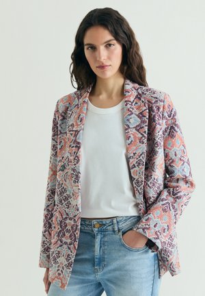 Mujer con blazer de estampado floral multicolor sobre una camiseta blanca y jeans azul claro, de pie con una mano en el bolsillo.