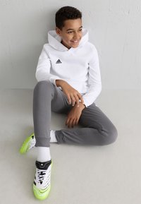 Hvid hætte sweatshirt med sort logo, grå joggingbukser og sorte samt neon grønne sportsko, siddende på en lys overflade.